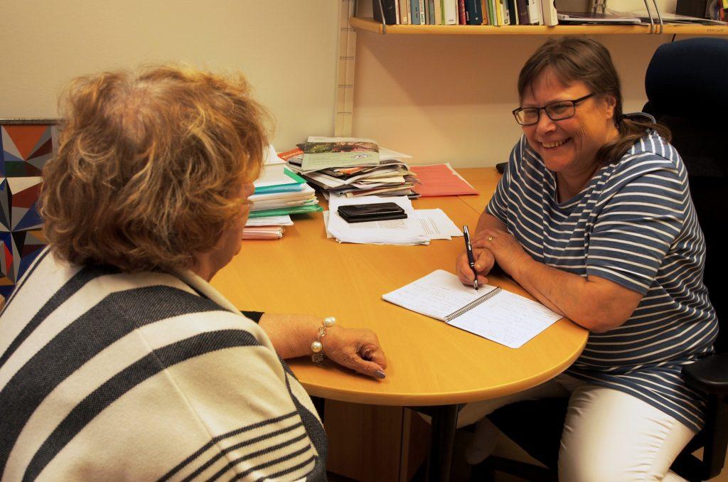 Aluepalvelut tukee asiakkaita kuulonäkövammaan liittyvässä palvelutarpeen selvittämisessä. Kuvassa asiakas keskustelee palveluasiantuntijan kanssa pöydän ääressä. Joskus arjessa on kommelluksia, joka naurattaa molempia.