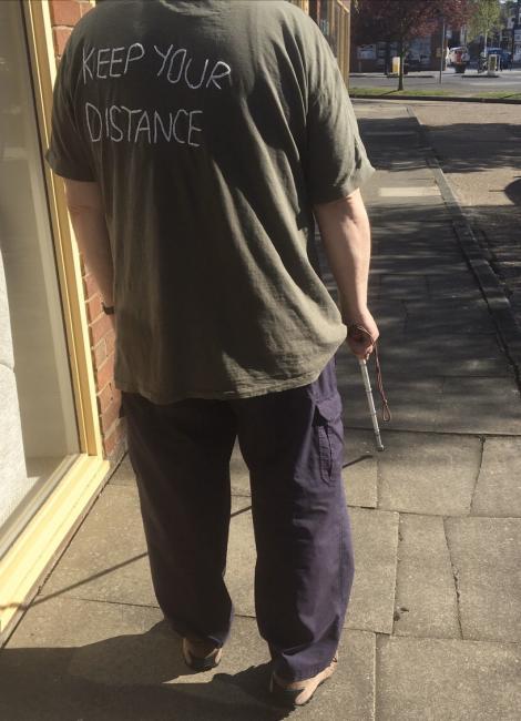 Miehellä on yllään t-paita, jossa lukee "Pidä etäisyyttäsi" Mies on kameraan selin päin ja hänestä näkyy kuvassa vartalo hartioista alaspäin. Miehellä on myös tumnat housut.