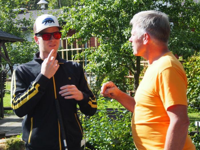Aarne Pirkola viittomassa Risto Hoikkaselle. Aarnella on lippalakki, aurinkolasit ja tumma verkkaritakki. Riston t-paita on keltainen. He ovat ulkona puutarhassa.