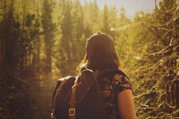 Nainen vaeltaa aurinkoisessa metsässä selkä kameraan päin. Hän kantaa selkäreppua. Hänestä näkyy vartalon yläosa.