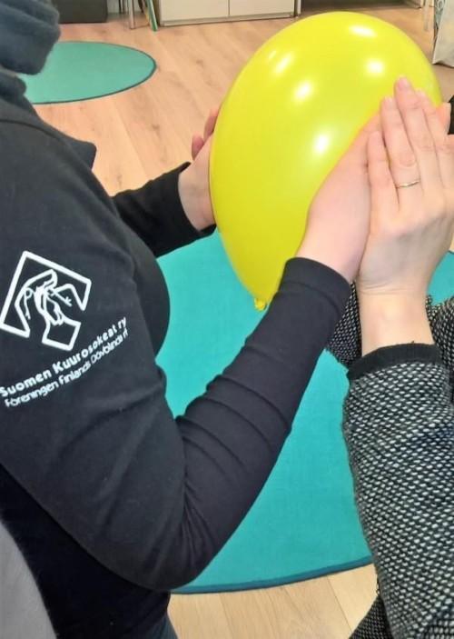 Kaksi henkilöä tunnustelee yhdessä keltaista ilmapalloa. Toisella henkilöllä on paidassa Suomen Kuurosokeat ry:n logo. Henkilöistä näkyy kädet ja vähän paitaa.