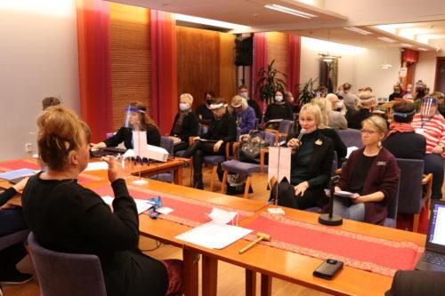 Yleiskuva Suomen Kuurosokeat ry:n syyskokouksesta 2020 Tampereelta. Etummaisena on kokouksen puheenjohtaja kuvattuna takaapäin pöydän ääressä. Kokouksen osallistujat istuvat penkeillä ja seuraavat kokousta. Yhdellä henkilöllä on kädessä mikrofoni ja hän puhuu.