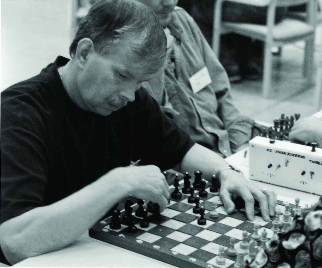 Mies istuu pelaamassa shakkia pöydän ääressä. Hän on siirtämässä mustia nappuloita shakkilaudalla. Hänen vieressään näkyy osittain toista henkilöä. Henkilöllä on nimikyltti paidassa.