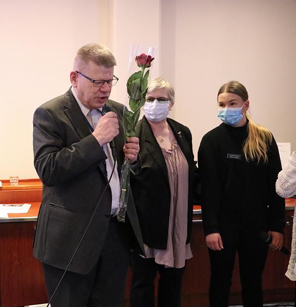 Timo Lehtonen pitämässä puhetta kokouksessa. Toisessa kädessä hänlellä punainen ruusu, joka kääritty läpinäkyvään sellofaaniin. Taustalla seisoo Tuula Hartikainen ja tulkki.