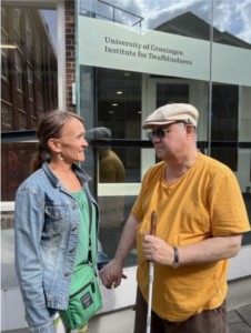 Russ Palmer ja Marja Cantell syyskuussa 2022 Groningenissa. Kuvailu: Russ on kuvassa oikealla. Hänellä on lippahattu, tumat aurinkolasit, oranssi t-paita sekä kädessään hänellä valkoinen keppi. Marja on Russin vierellä ja katsoo häntä. Marjalla on auki oleva farkkutakki ja t-paita. Olkapäiltä roikkuu kaulalaukku. 