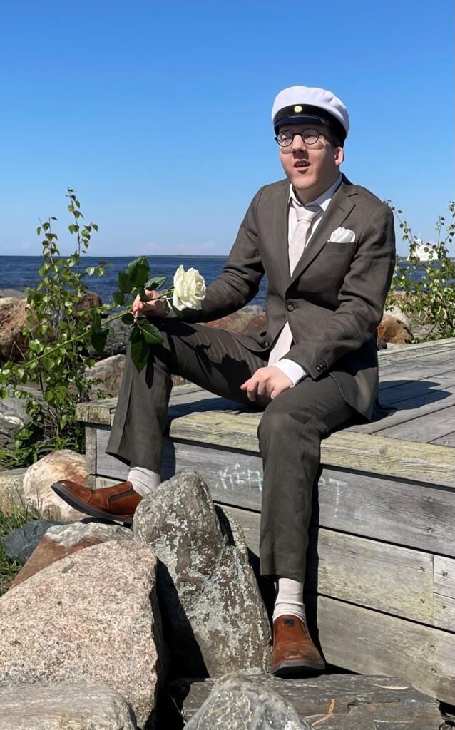 Lauri kuvattu edestä. Hänellä on yllään puku, valkoinen kauluspaita, kravatti ja päässään ylioppilaslakki. Puvun taskussa on liina. Laurin kädessä on valkoinen ruusu. Taustalla näkyy vesistöä.