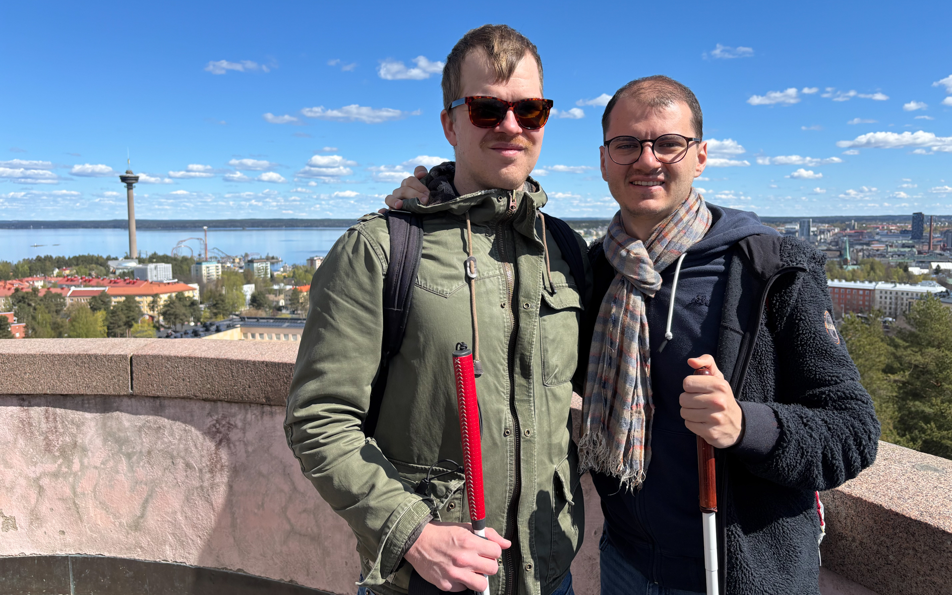Aarne ja Marco kuvattuna edestä. Heillä molemmilla on käsissään valkoinen keppi. Aarnella on tummat aurinkolasit, Marcolla silmälasit. Molemmilla on päällään takki. Taustalla avautuu järvimaisema ja Näsinneula. Taivas on aurinkoinen.