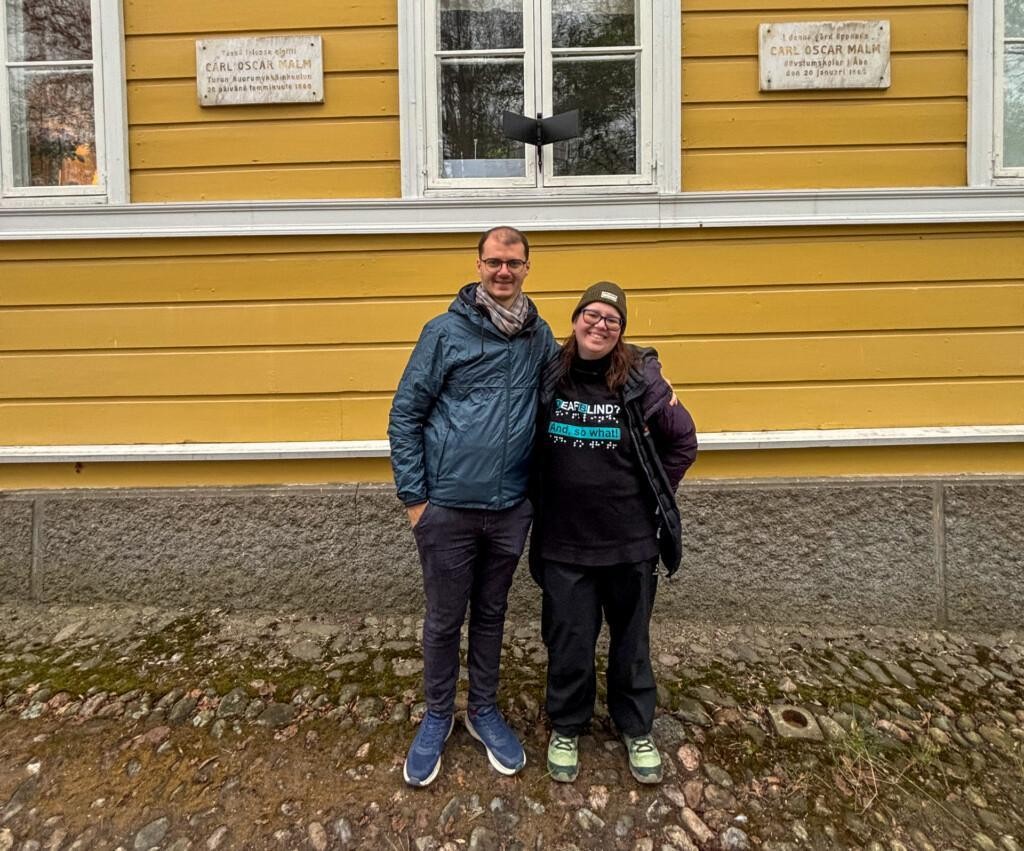 Marco ja Roberta seisovat koulurakennuksen edessä. Marco pitää kättään Robertan olkapäällä. Marcolla on takki, kaulahuivi ja silmälasit. Robertalla on hattu, silmälasit, takki avoinna ja paita, jossa on teksti englanniksi ja pisteet: ”Deafblind…and so what!” Heidän takanaan seinällä vasemmalla on kyltti, jossa lukee ”Tässä talossa aloitti Carl Oscar Malm Turun kuuromykkäinkoulun 20. päivänä tammikuuta 1860”. Oikealla puolella olevassa kyltissä on sama teksti ruotsiksi. Keskellä seinää on ikkuna.