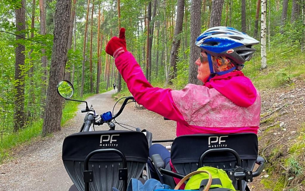 Anne istuu tandemin oikealla penillä. Hän pitää peukaloaan ylhäällä. Päässään hänellä on kypärä, kasvoillaan aurinkolasit ja yllään vaaleanpunainen takki. Edessä on hiekkatie ja puita.