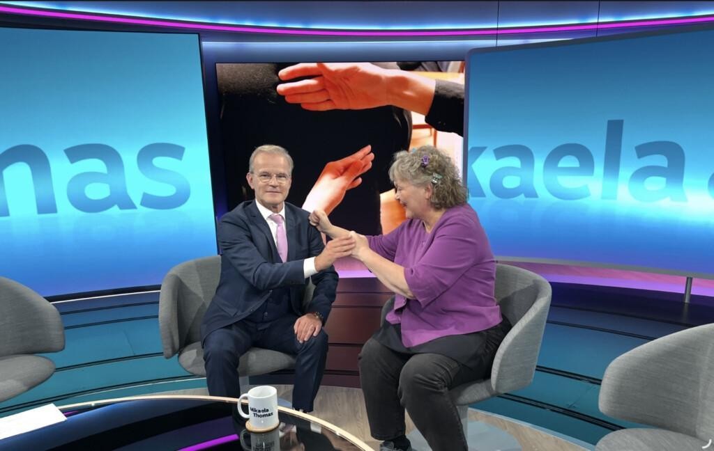 Riitta Lahtinen ja Thomas Sandholm istuvat tuoleilla. Riitta esittelee kahvi-haptiisia Thomakselle olkapäähän. Hän vastaa Riitalle kyllä-palautteella. Taustalla näkyy studion taustaa ja kuvituskuva kahdesta kädestä selkää vasten.