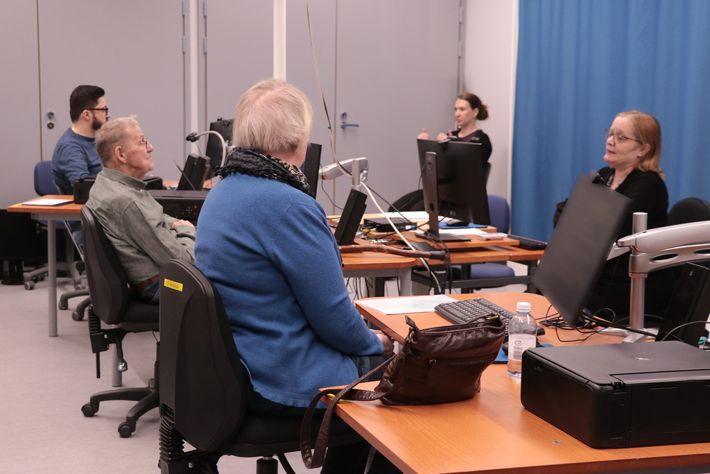 Henkilöitä ICT-koulutusluokassa.  He istuvat tuoleilla, edessä on tietokoneita. Ohjaaja istuu kuvan oikeassa reunassa tietokoneen ääressä. Taustalla viittomakielen tulkkausta.