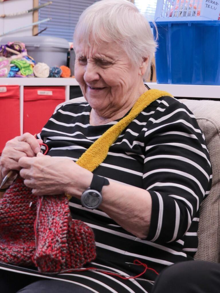 Hilkka kutoo. Kädessään hänellä on kutimet ja neule. Viistosti takana näkyy laatikoita ja tavaroita. Hilkalla on tummaraitainen paita ja lyhyet vaaleat hiukset.