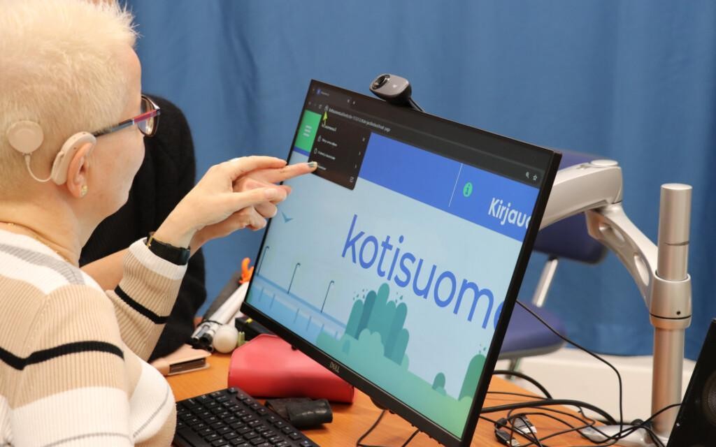 Henkilö istuu tietokoneen ääressä. Toinen henkilö on hänen vieressään ja ohjaa henkilön kättä tietokoneen ruudulla. Pöydällä on näyttö ja näppäimistö.