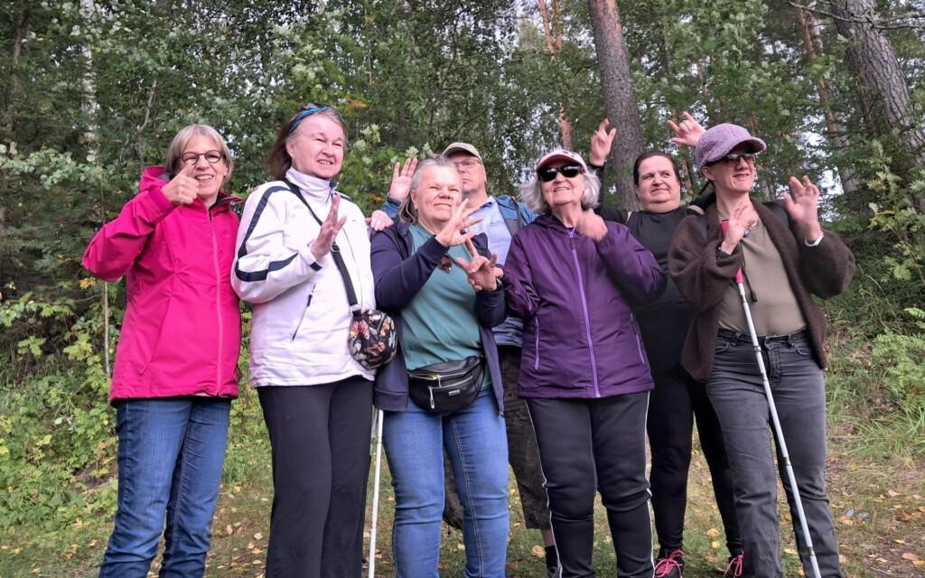 Henkilöt seisovat ryhmäkuvassa vierekkäin ulkona. He viittovat käsillään. Osa katsoo kohti kameraa, osa kamerasta sivuun. Taustalla on metsää.