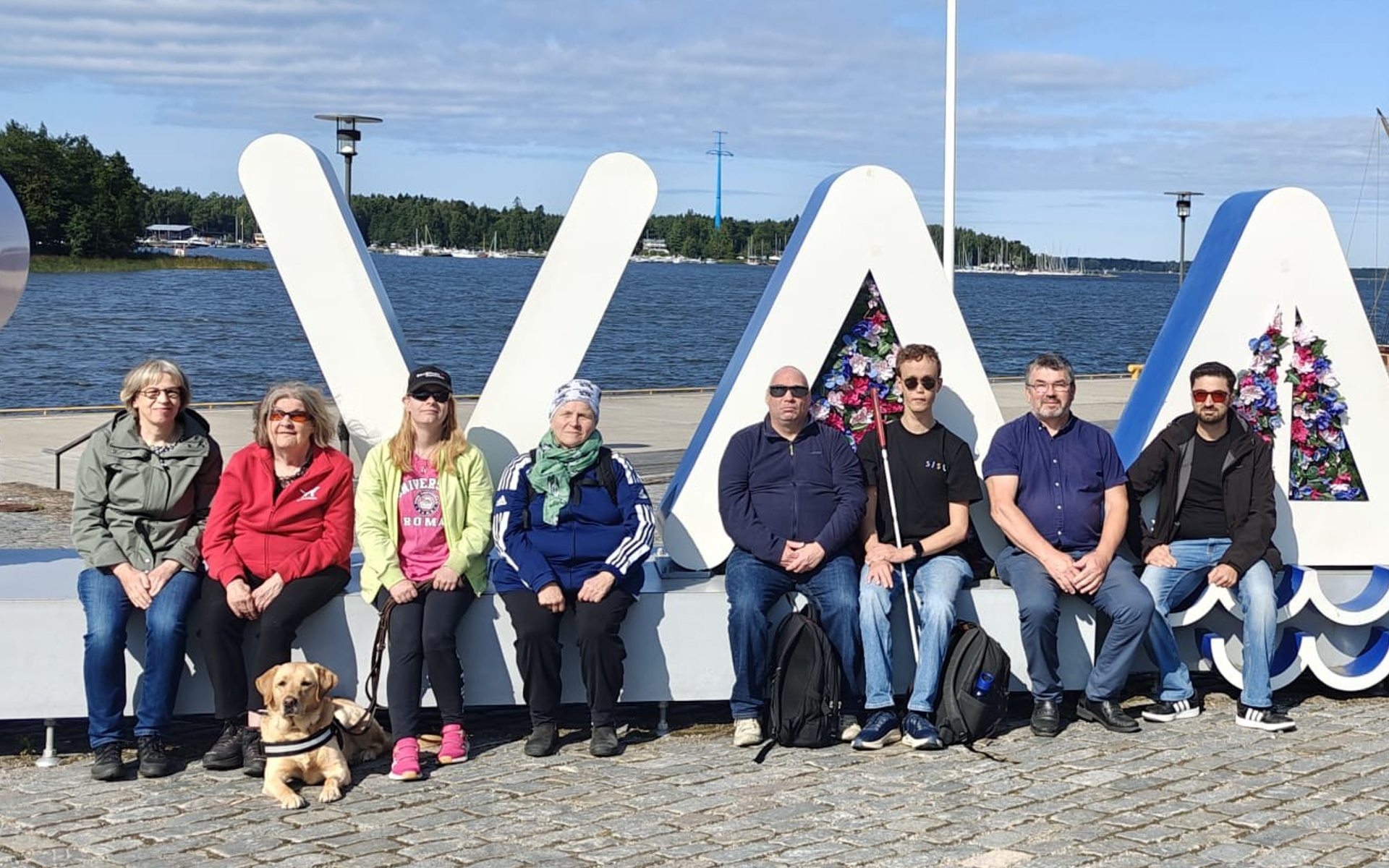 Henkilöitä ryhmäkuvassa ulkona. He istuvat vierekkäin, taustalla näkyy osittain isolla tekstillä: ”VAASA”. Usealla henkilöllä on kasvoillaan aurinkolasit. Yhdellä henkilöllä on vieressään opaskoira, yksi henkilö pitää kädessään valkoista keppiä. Keli on poutainen.