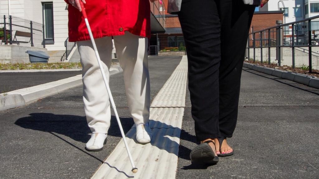 Kahden henkilön jalat. Vasemmalla henkilöllä näkyy valkoinen keppi, joka seuraa opastuslaattaa.
